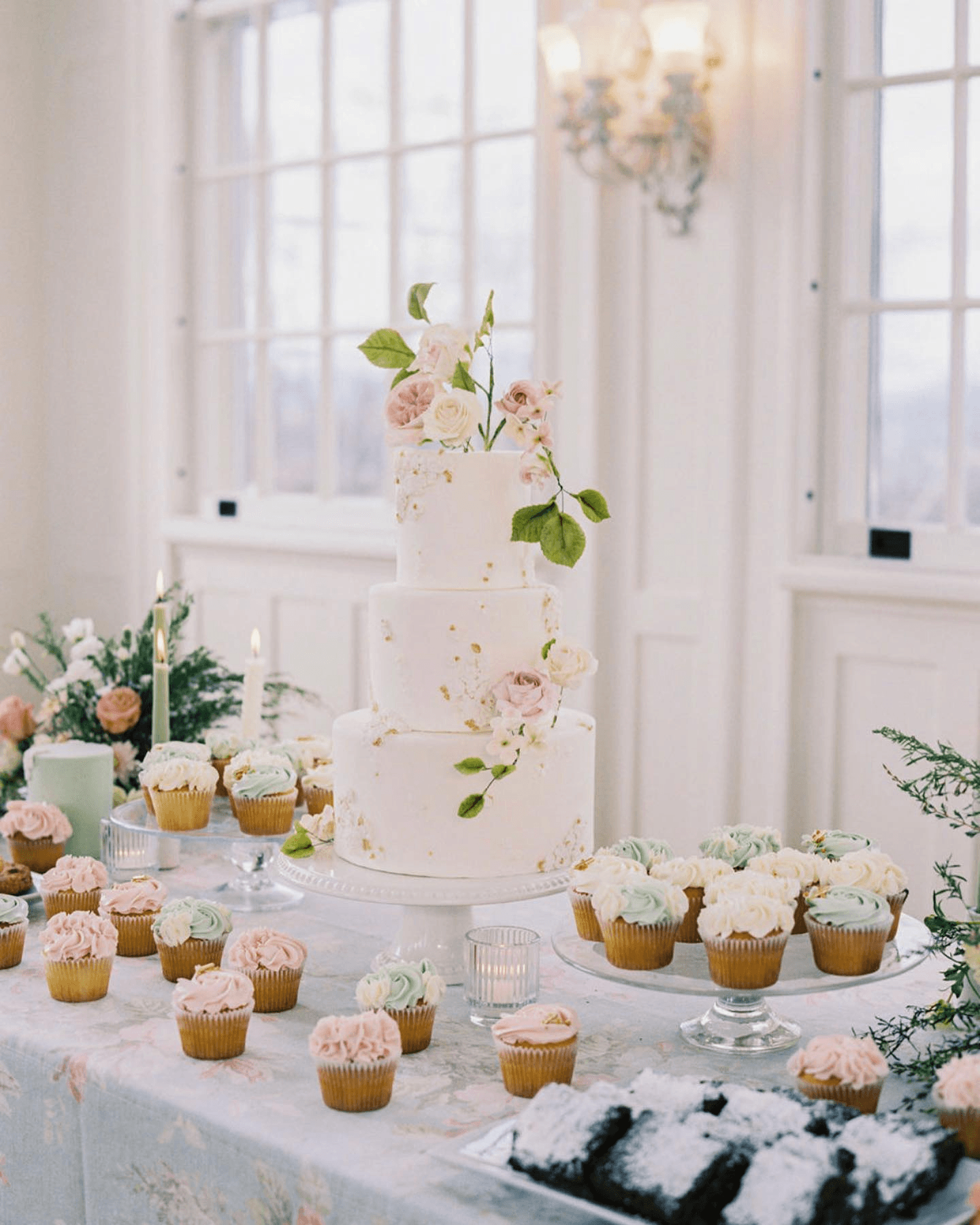 A beautifully decorated three-tiered wedding cake with floral accents, surrounded by an array of colorful cupcakes and candles on a floral tablecloth.